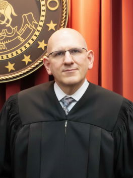 Headshot of Justice Carlos Samour in judge's robe in front of red curtains and state seal