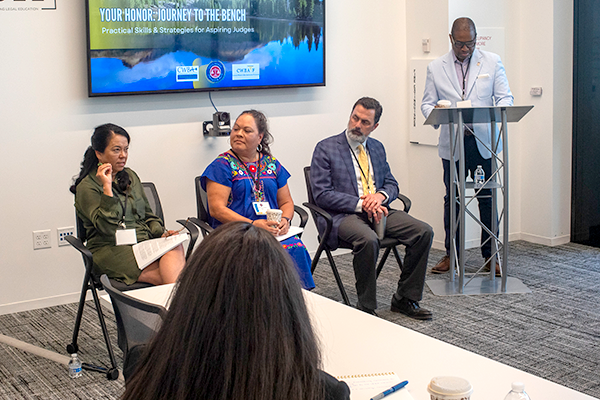 A panel of 3 guests sitting with one lady in a green dress speaking. The group is looking at her at she speaks. An African American man is standing at a podium next to the seated panel.