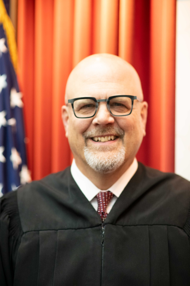Justice William Hood with glasses and a short gray beard wears a black judicial robe over a white shirt and red patterned tie, standing in front of red curtains and a partial view of the American flag.